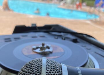Close-up of a microphone and DJ turntable in focus with a blurred outdoor pool in the background, featuring swimmers and a red lifeguard umbrella under a sunny sky.
