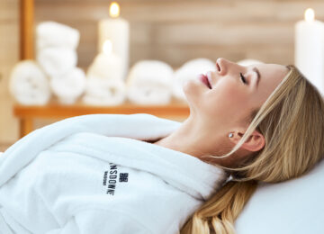 Woman relaxing in a white spa robe with candles and towels in the background, waiting for a facial.